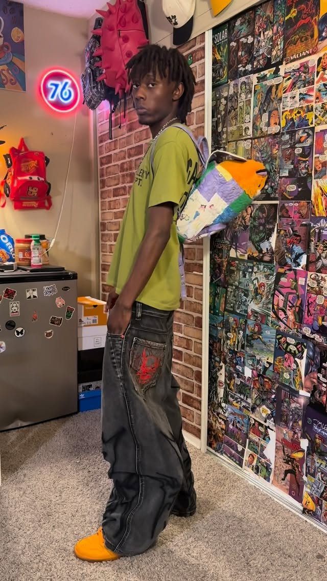 Man wearing a green t-shirt, baggy dark jeans, and orange shoes standing in a room.