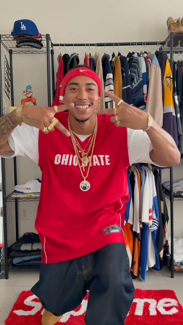 A man smiling and posing in a red basketball jersey, gold jewelry, and a red headband.
