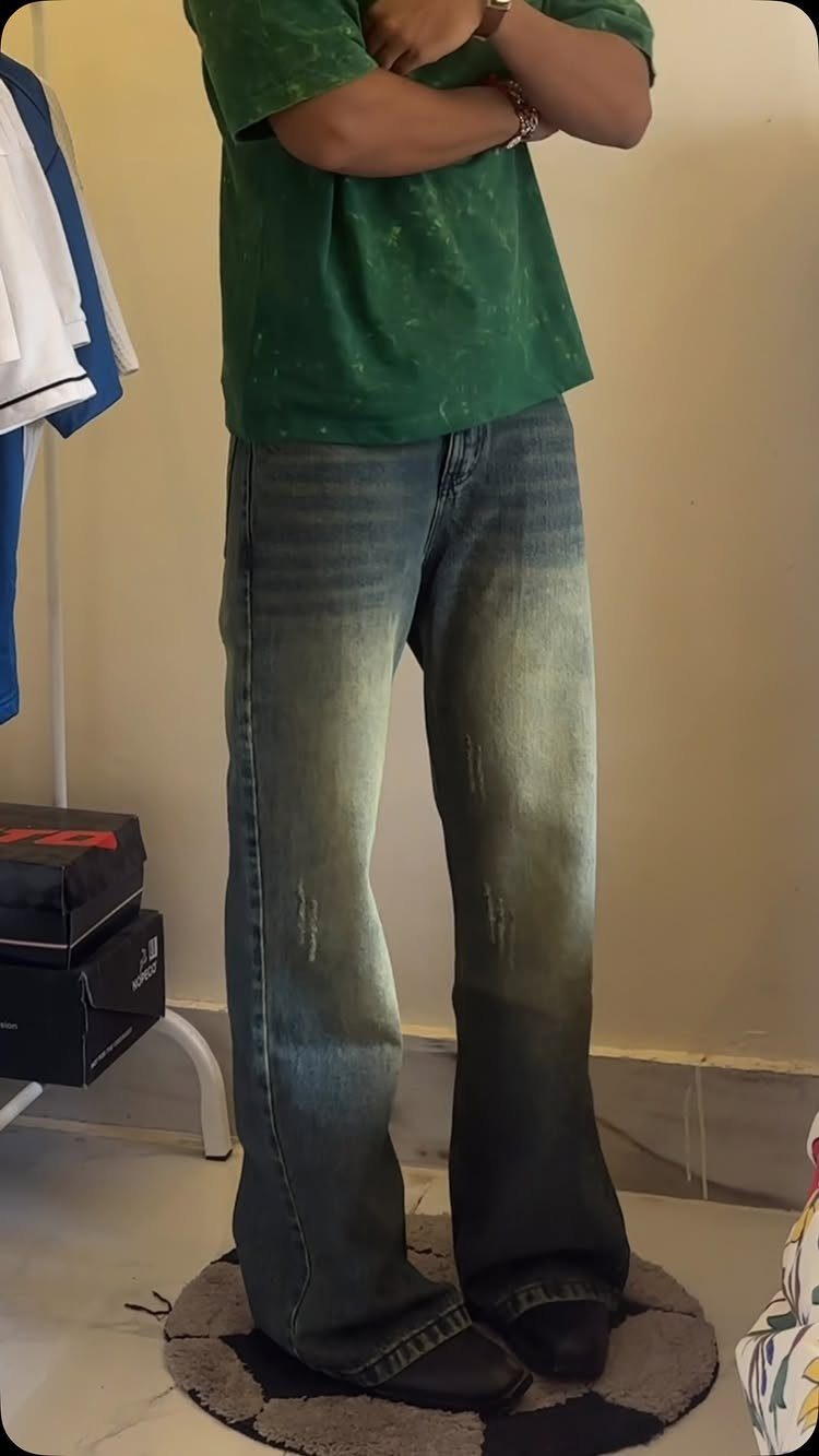 Man wearing green t-shirt and wide-leg boot-cut denim jeans standing indoors.