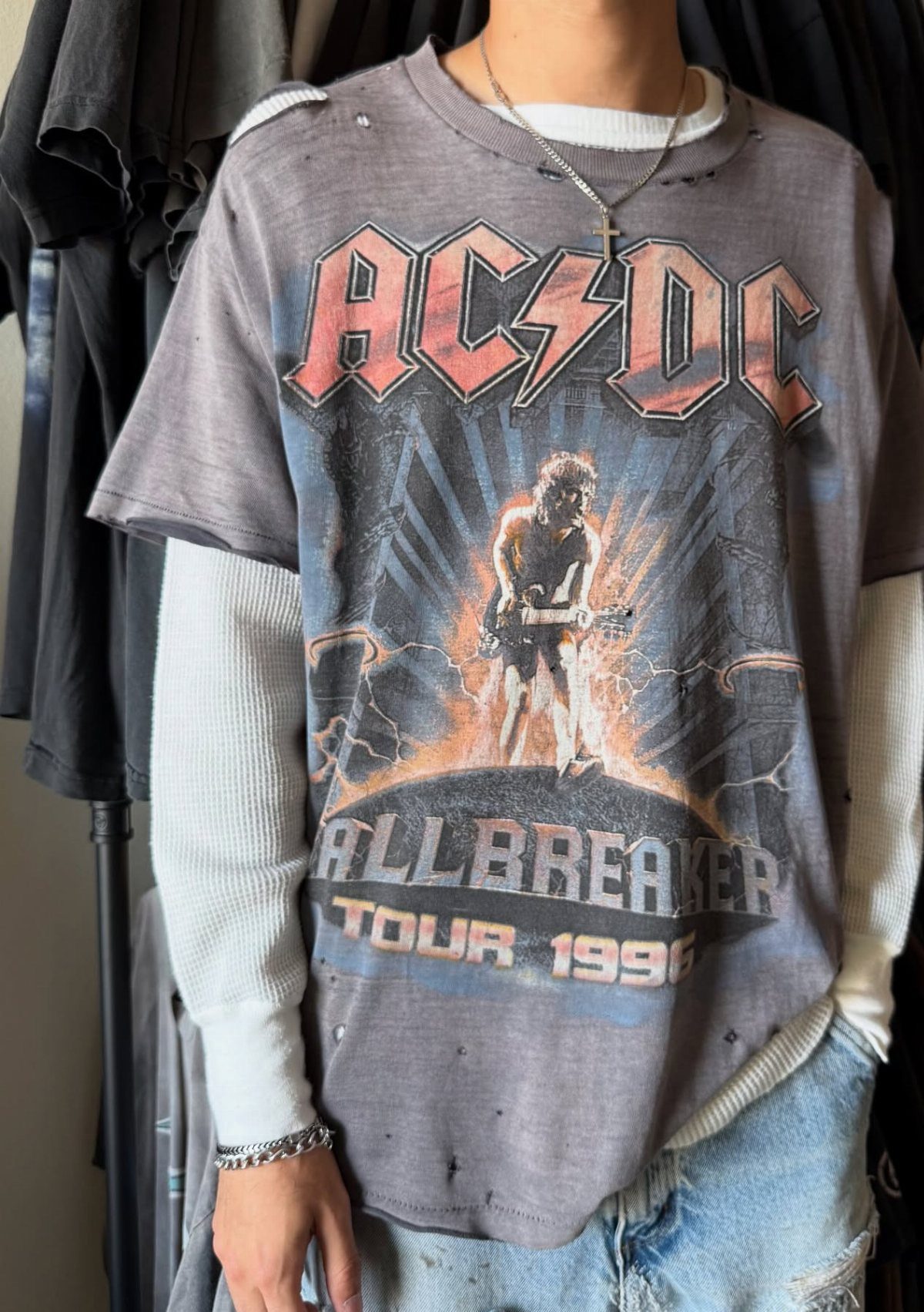 A person wearing a distressed grey AC/DC graphic t-shirt over a white long-sleeved shirt and blue jeans.