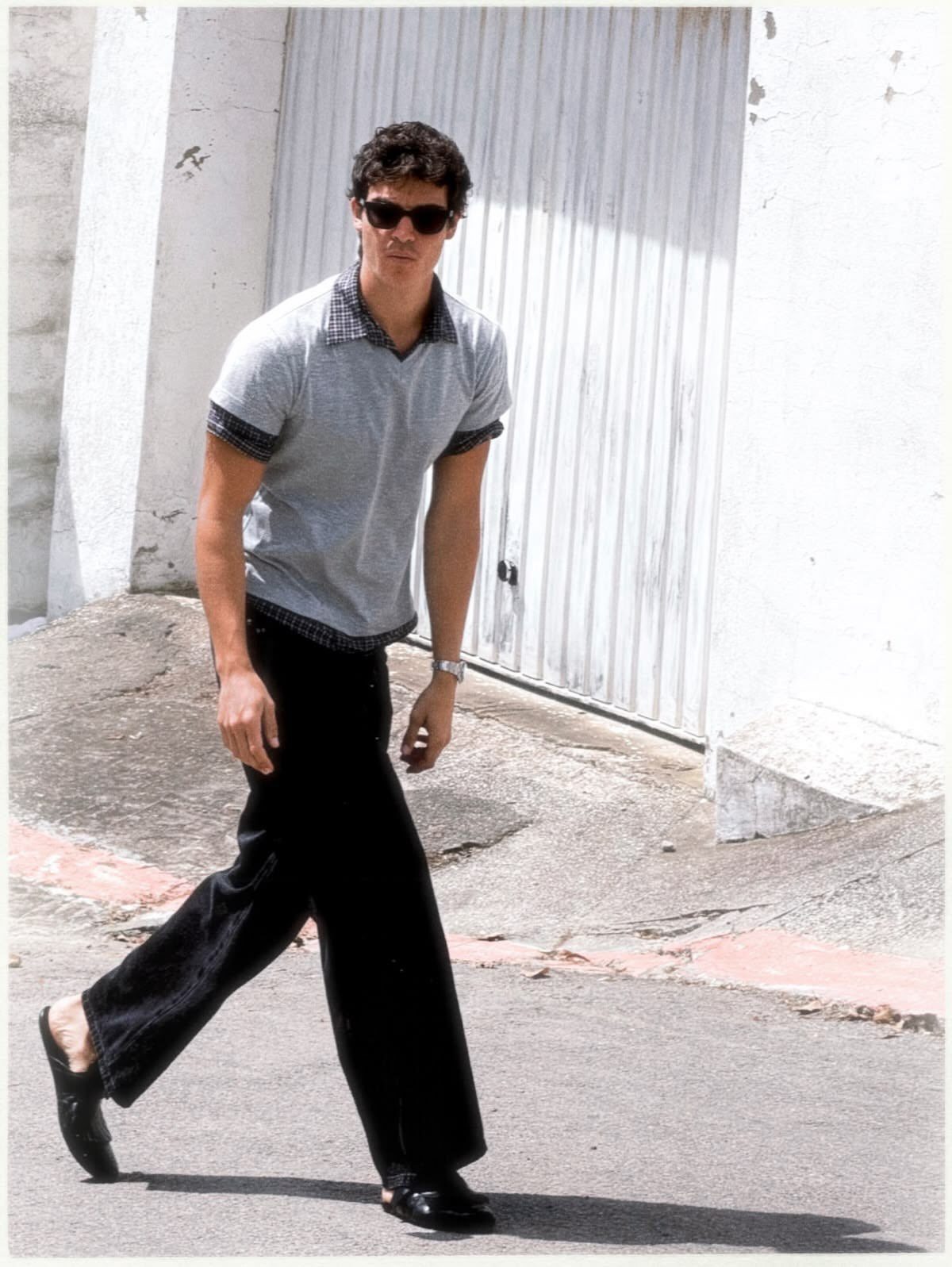 A man in sunglasses wearing a grey polo shirt and black flared trousers walking outdoors.
