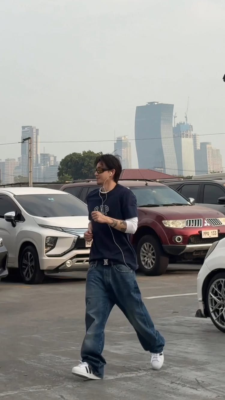 A man walking in a parking lot wearing a navy t-shirt, white long-sleeve shirt, and baggy blue jeans.