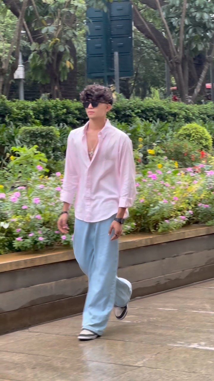 A man in a pink shirt and baggy blue trousers walking past a flower garden.