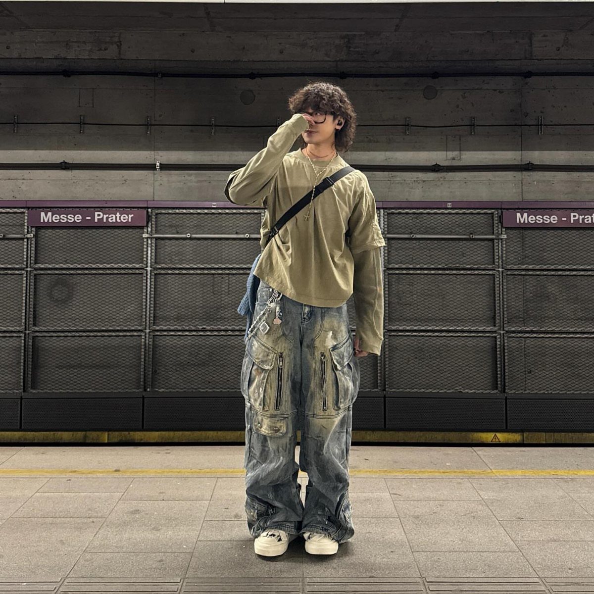 A person wearing oversized olive shirt and baggy distressed denim cargo pants standing in a subway station.