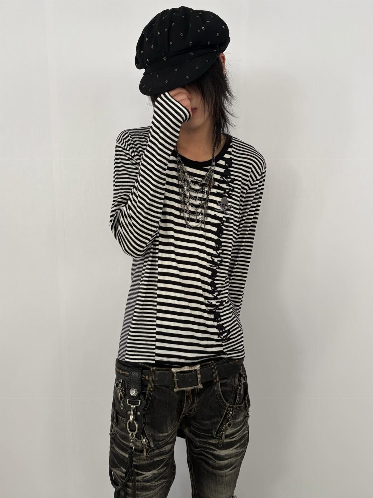 Person wearing a striped shirt, distressed jeans, layered necklaces, and a black newsboy cap.