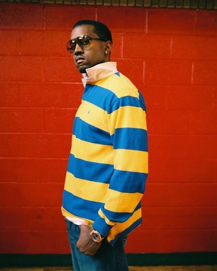 A man in a blue and yellow striped shirt and sunglasses posing against a red wall.