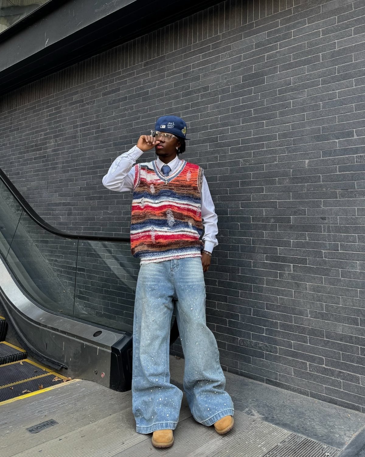 Man in a colorful knit vest and baggy denim jeans standing near an escalator.