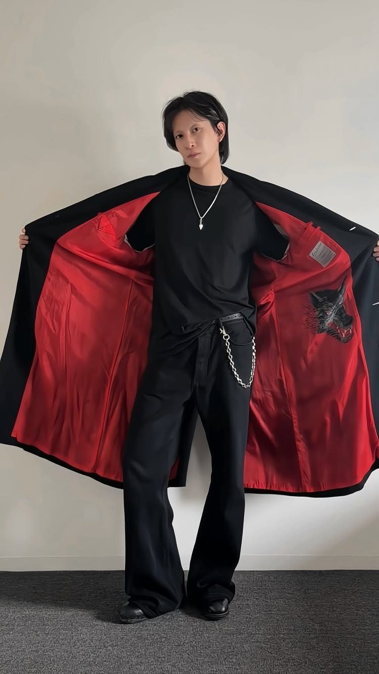 Man in black flared trousers and t-shirt holding open a black coat with red lining.