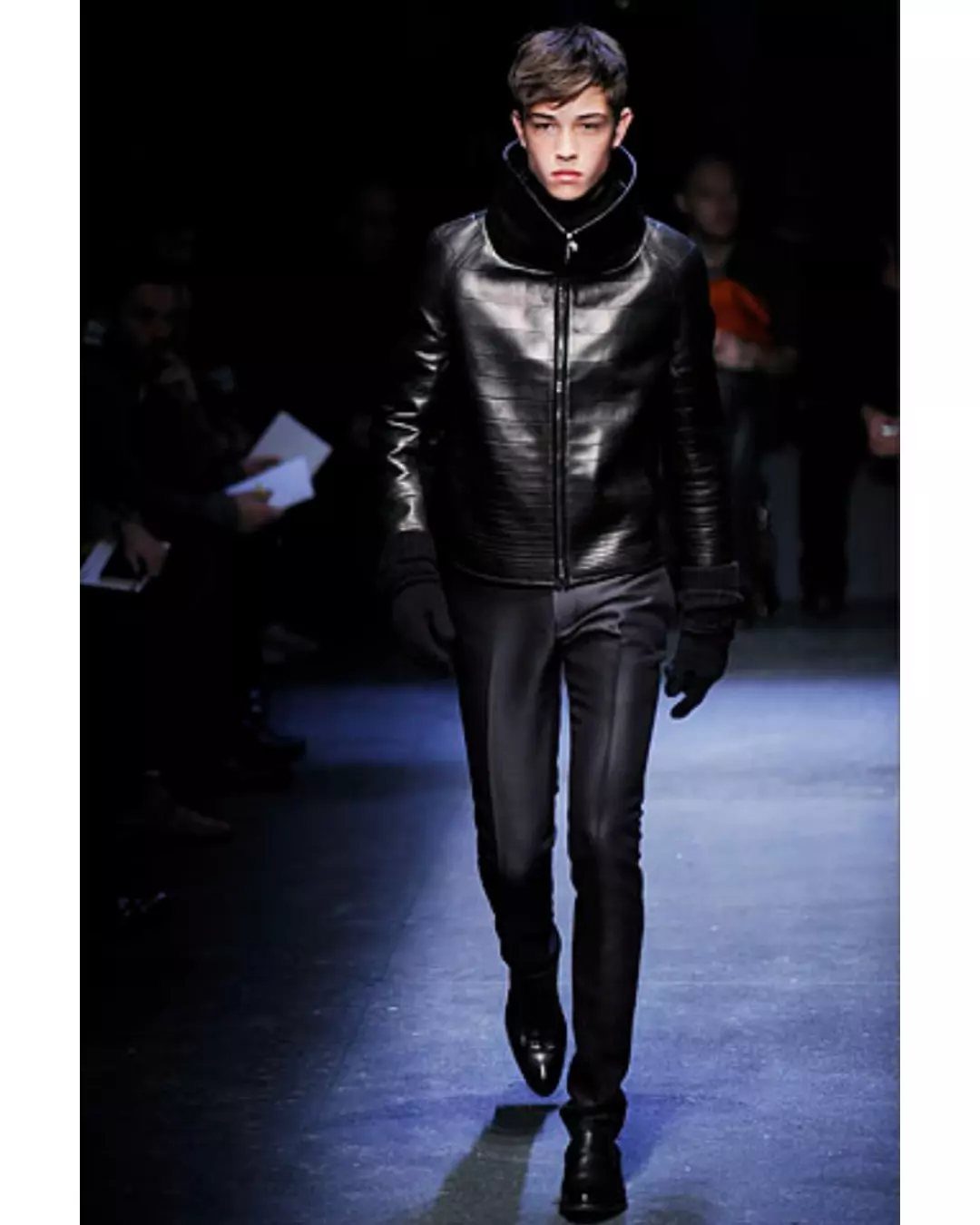 Male model walking a runway in a black leather jacket and slim trousers.