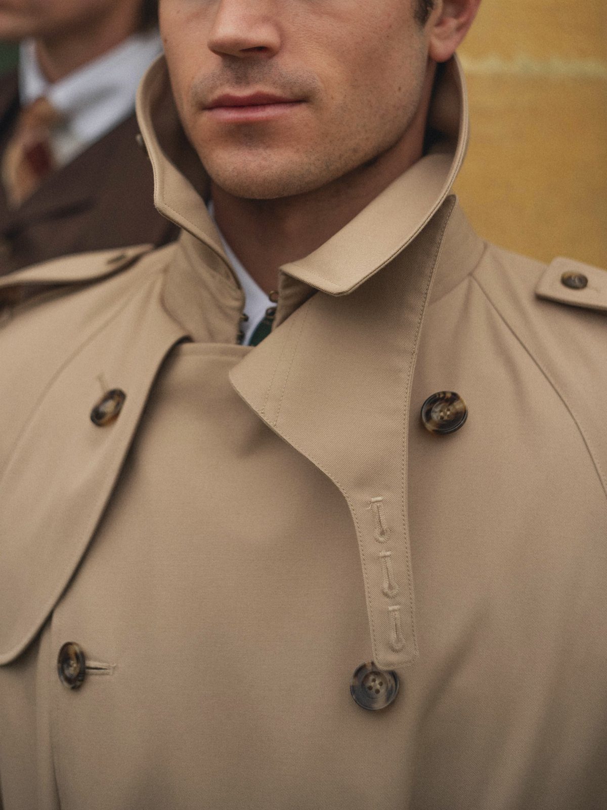 Close-up of a man wearing a tan trench coat with a high collar and dark buttons.