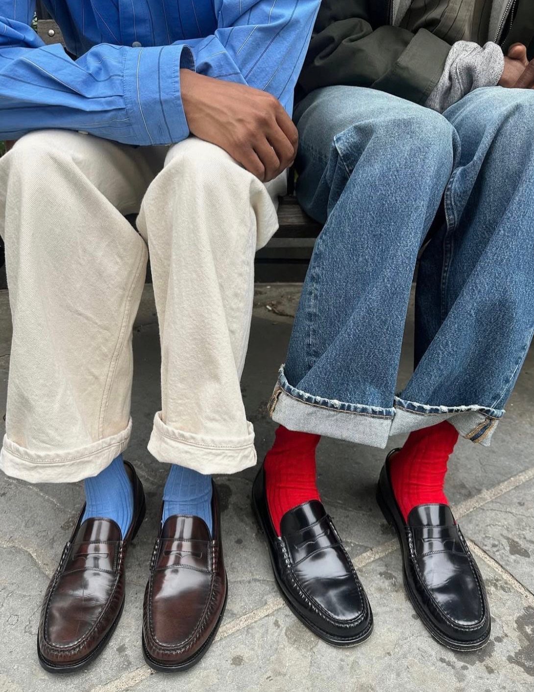 Two people sitting wearing various trousers, colorful socks, and leather loafers on a sidewalk.