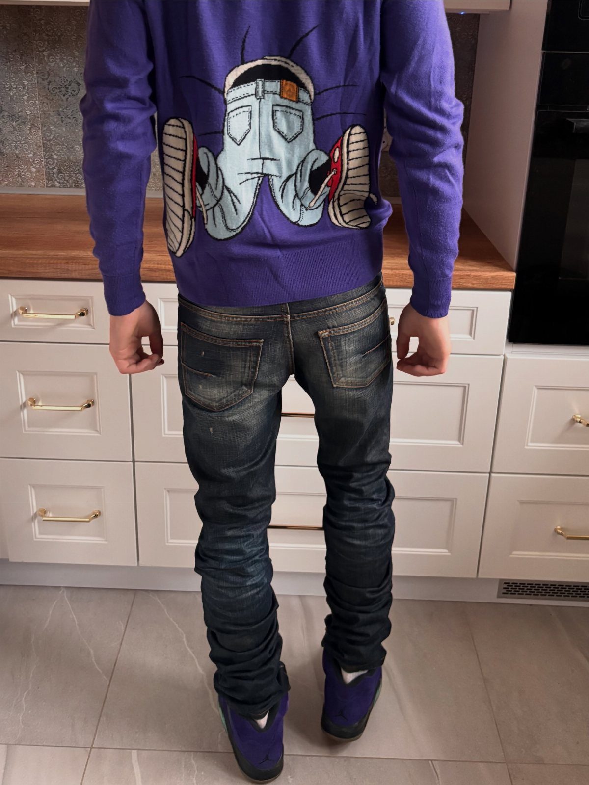 Man wearing a purple graphic sweater, dark slim jeans, and purple shoes standing in a kitchen.