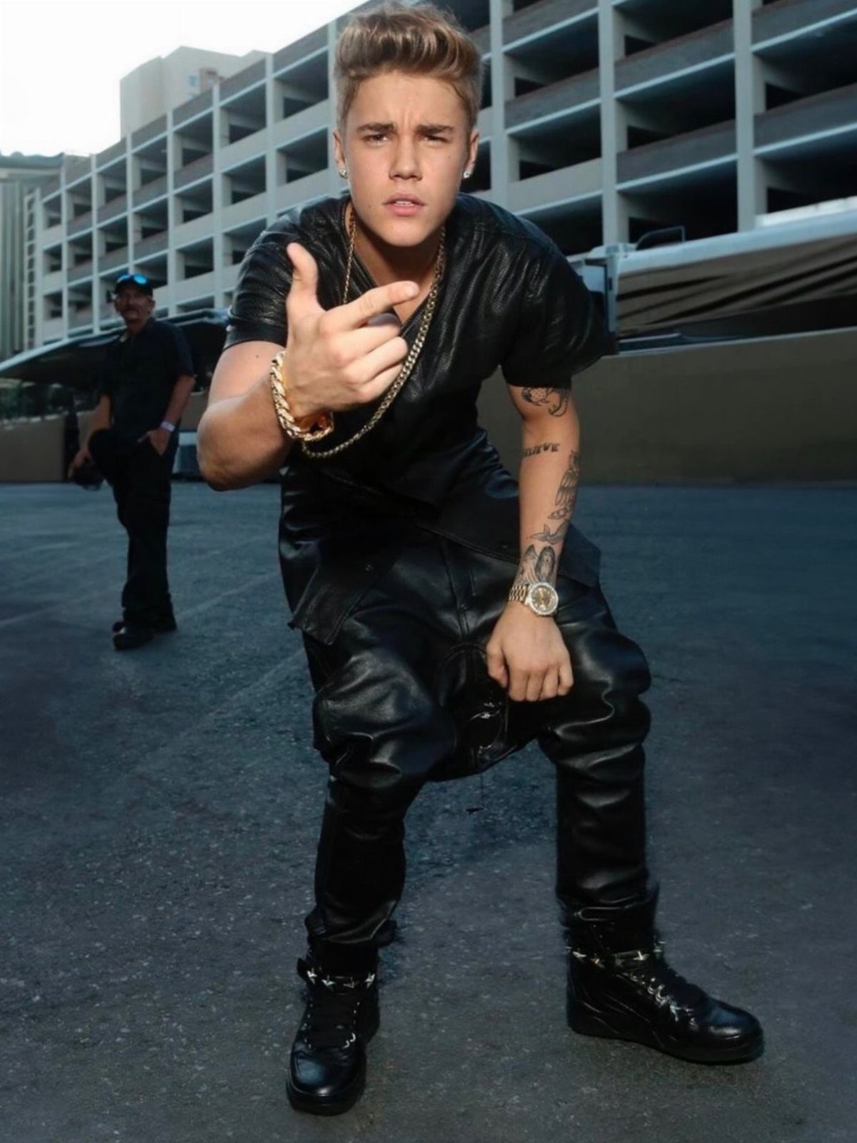 Young man in black leather clothing and gold jewelry posing in an urban outdoor setting.