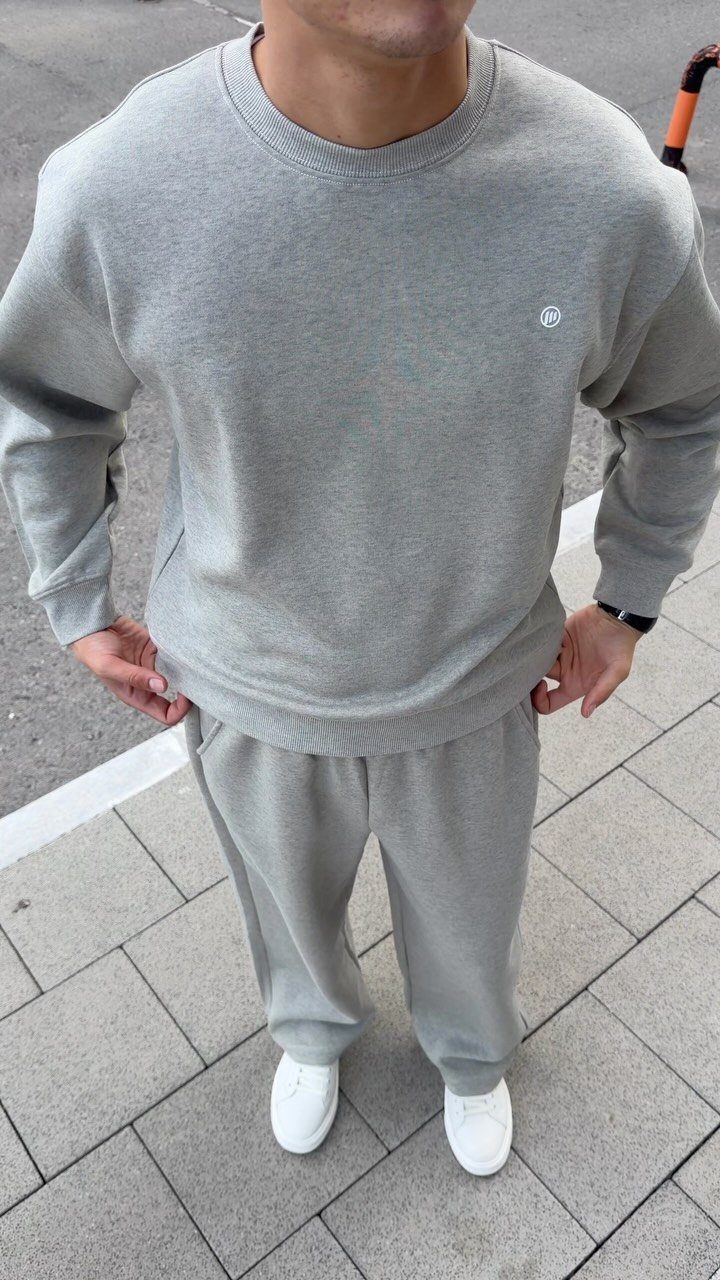 Man wearing a grey sweatshirt and matching sweatpants with white sneakers on a tiled sidewalk.