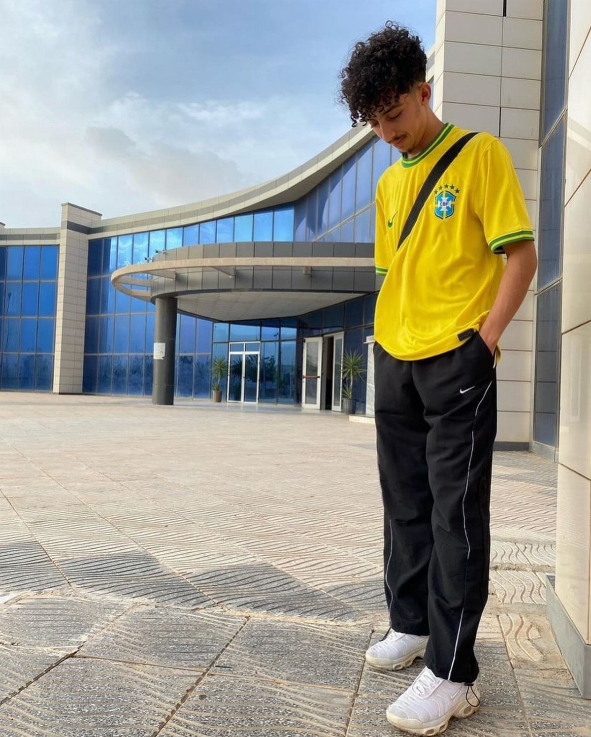 A young man wearing a yellow soccer jersey and black joggers standing in front of a glass building.