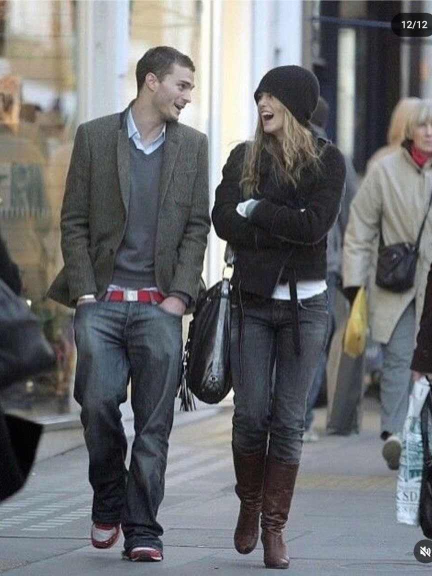 A man in a blazer and sweater walking alongside a woman in a black beanie and boots.