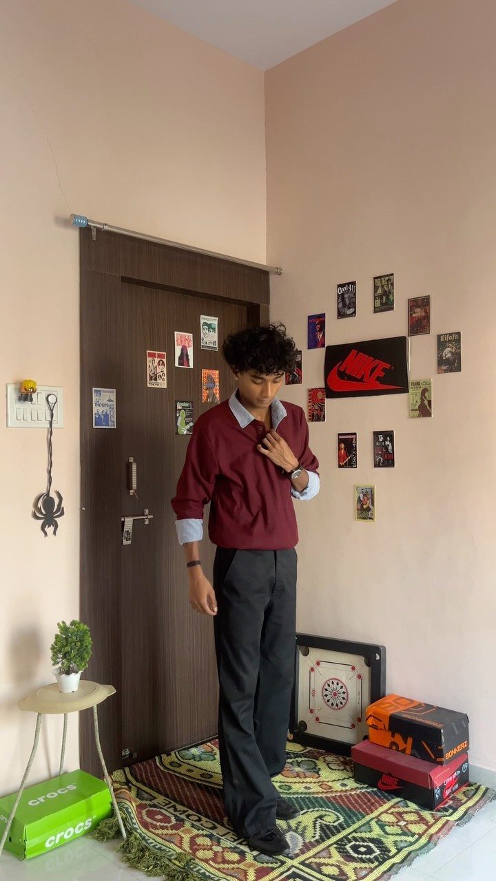 A young man wearing a maroon collared shirt layered over a light blue shirt and dark trousers.