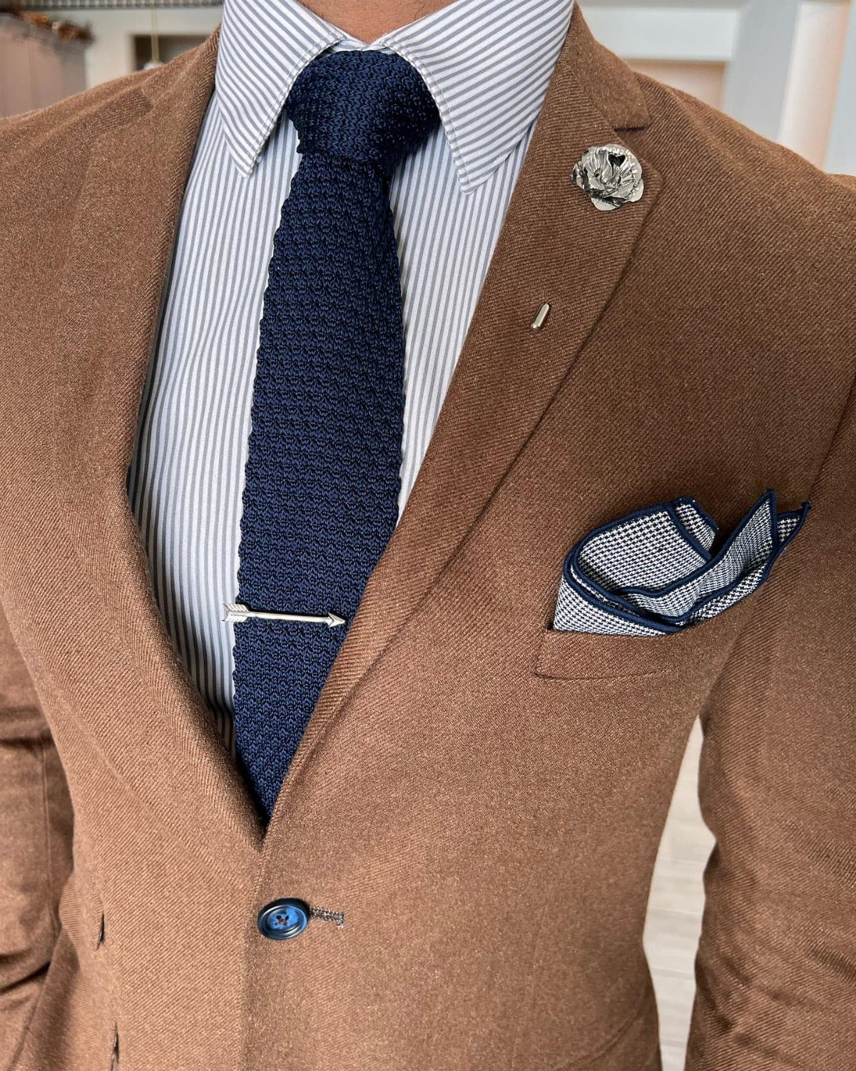 A person wearing a brown blazer, a striped shirt, and a navy blue knitted tie with a silver clip.