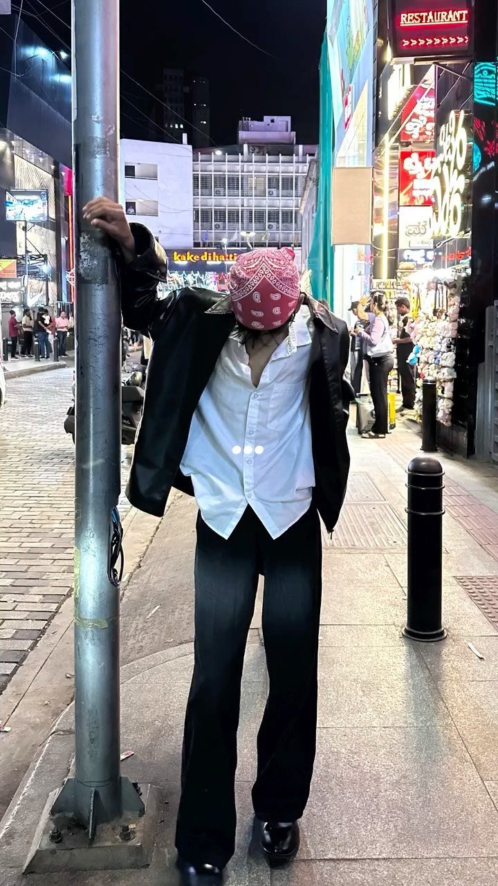 A person wearing a pink patterned bandana and black jacket leans against a pole at night.