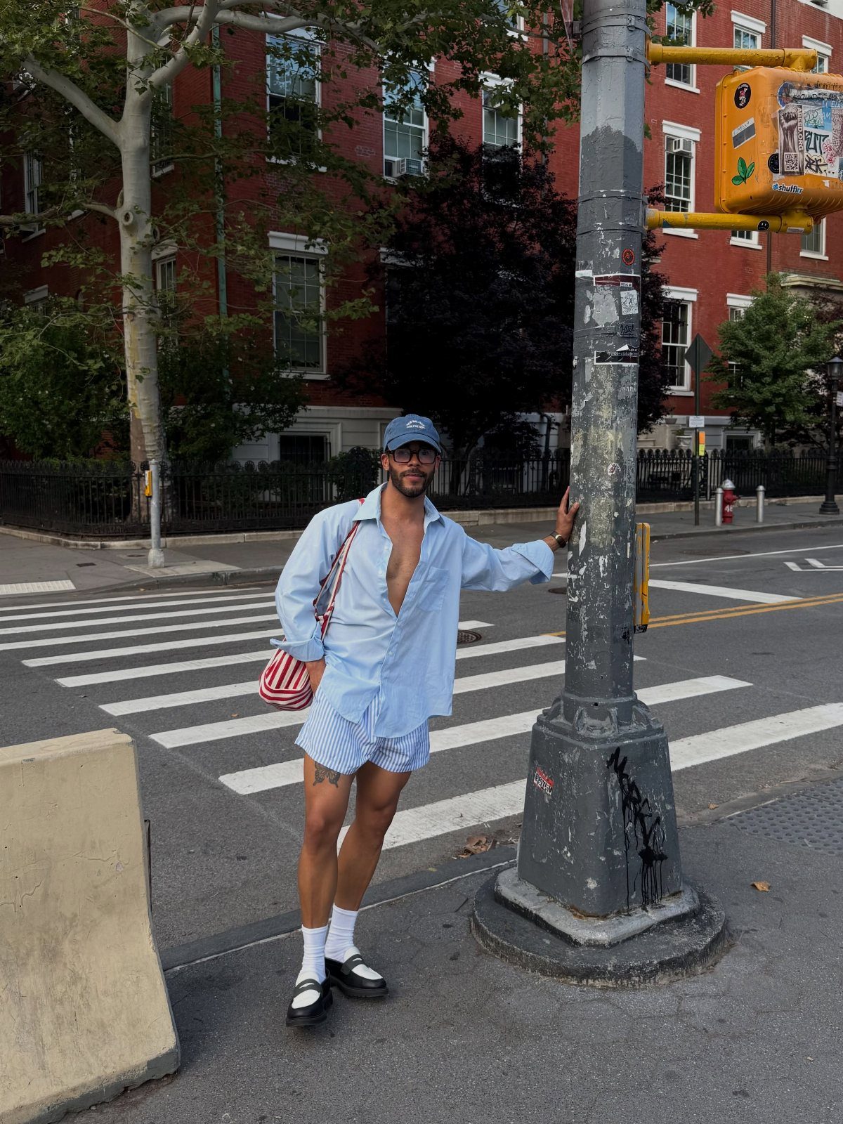 A man in light blue shorts and an unbuttoned shirt posing next to a street pole.