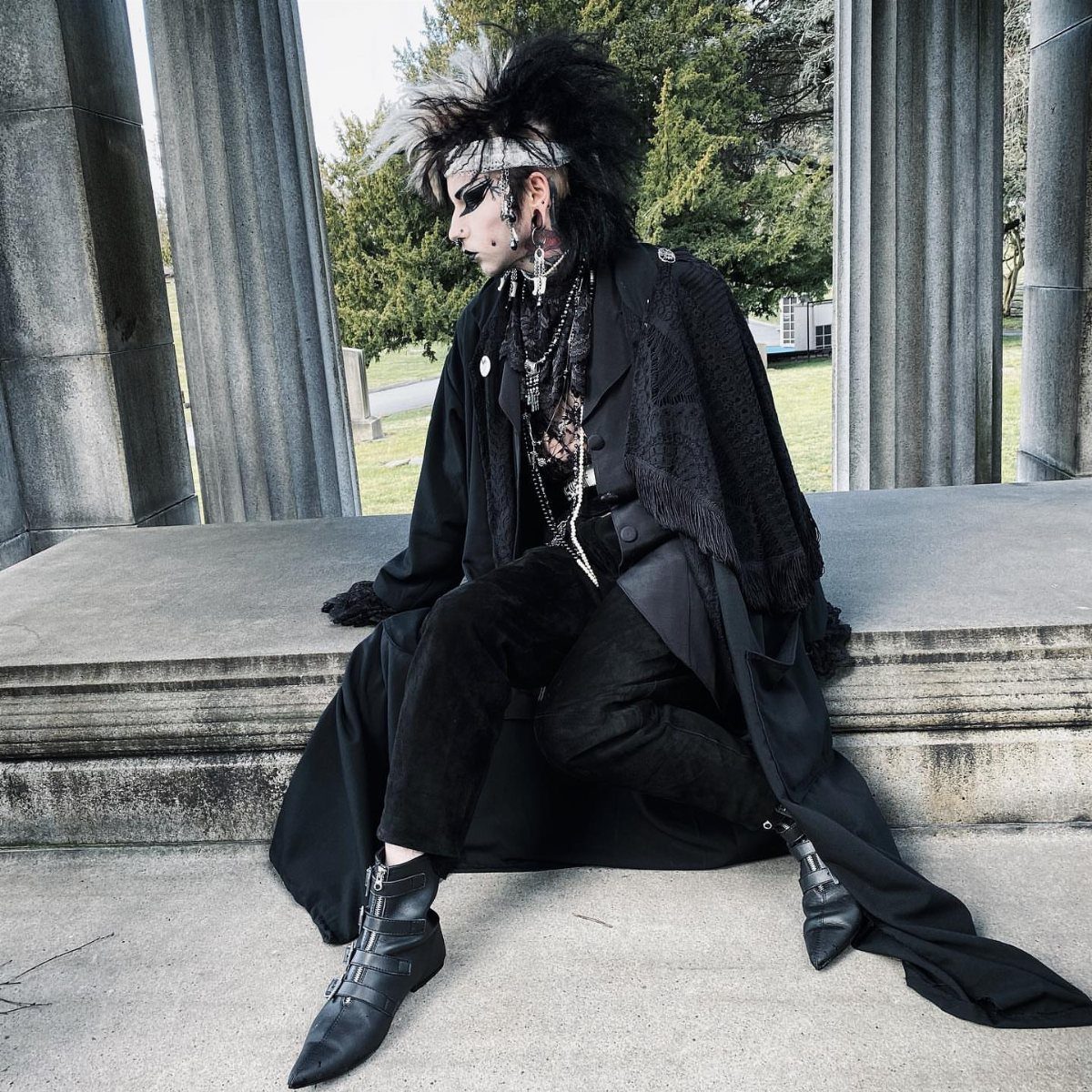 A person in an all-black goth outfit sits on stone steps near large columns.