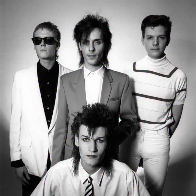 Four men with dark textured hair posing in black and white studio attire.