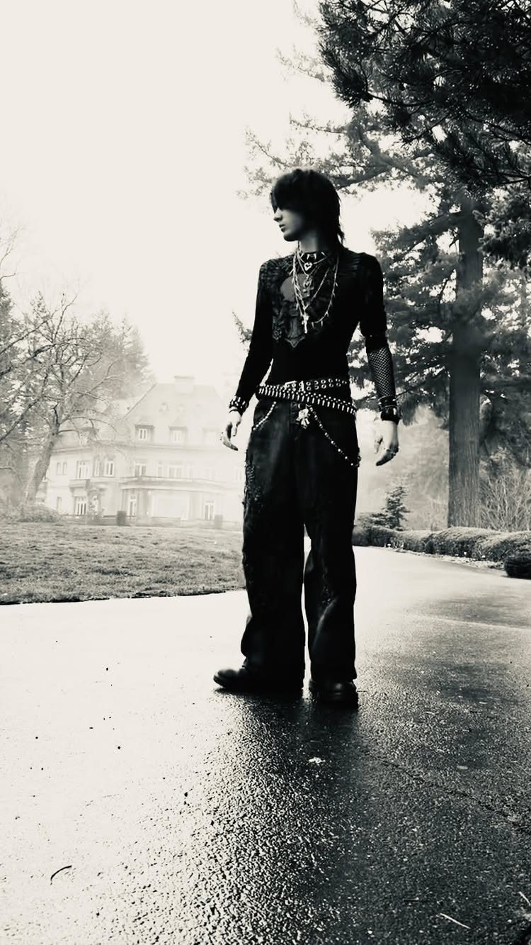 A person wearing a black gothic outfit with studded belts and necklaces stands in a misty park.