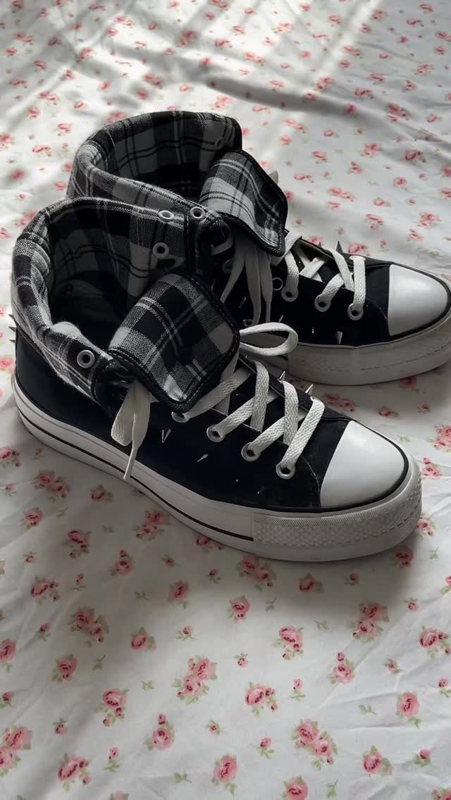 Black high-top sneakers with plaid tongues resting on a white floral patterned bedsheet.