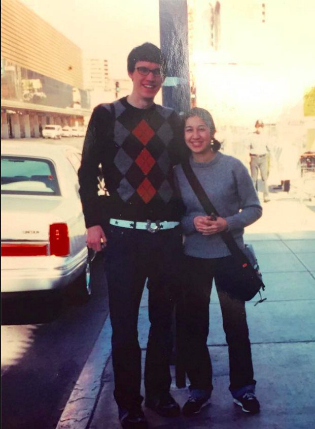 A man in an argyle sweater and a woman in a grey shirt posing on a city street.