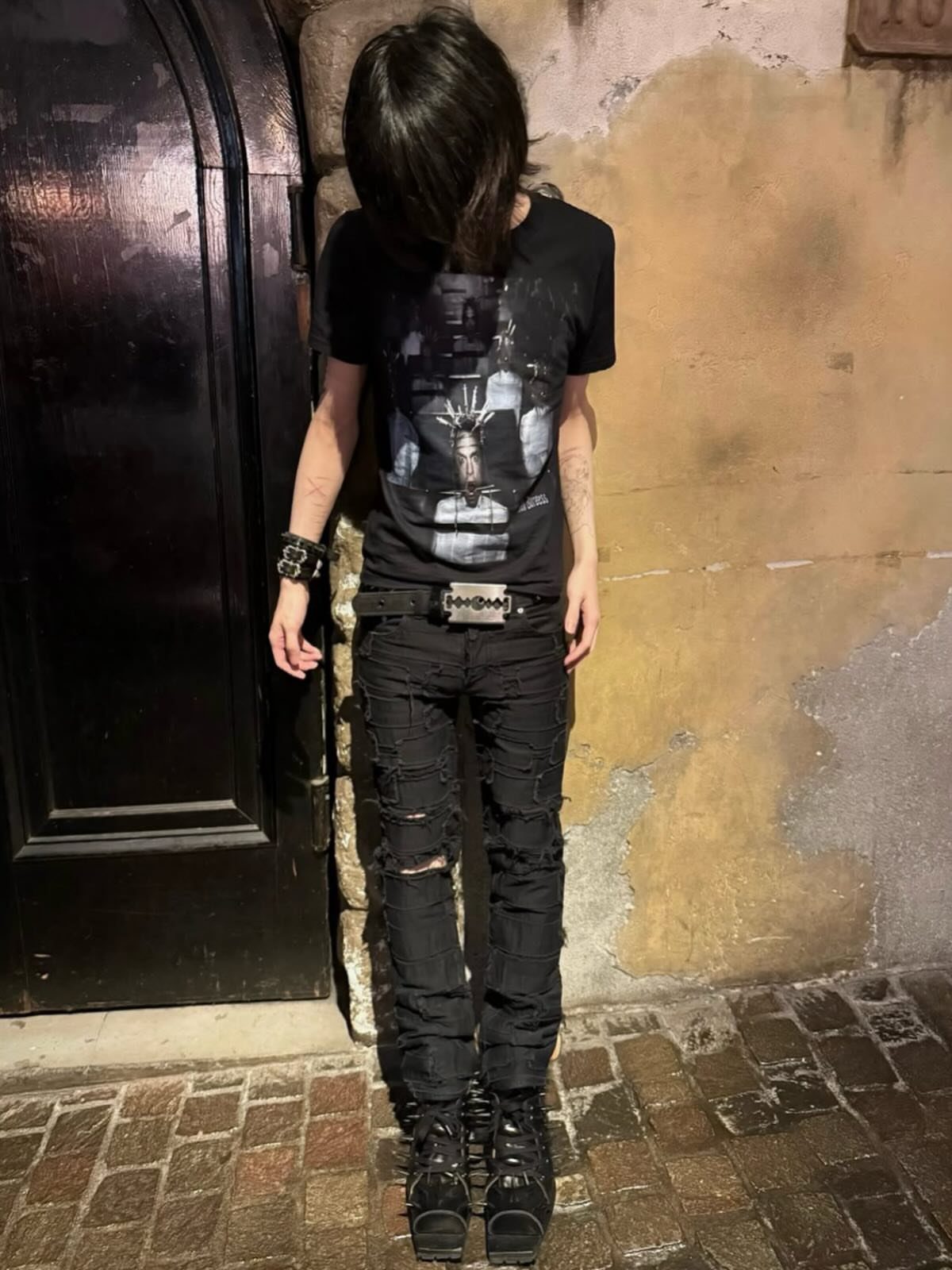 A person wearing black graphic t-shirt, studded belt, and ripped black distressed jeans.