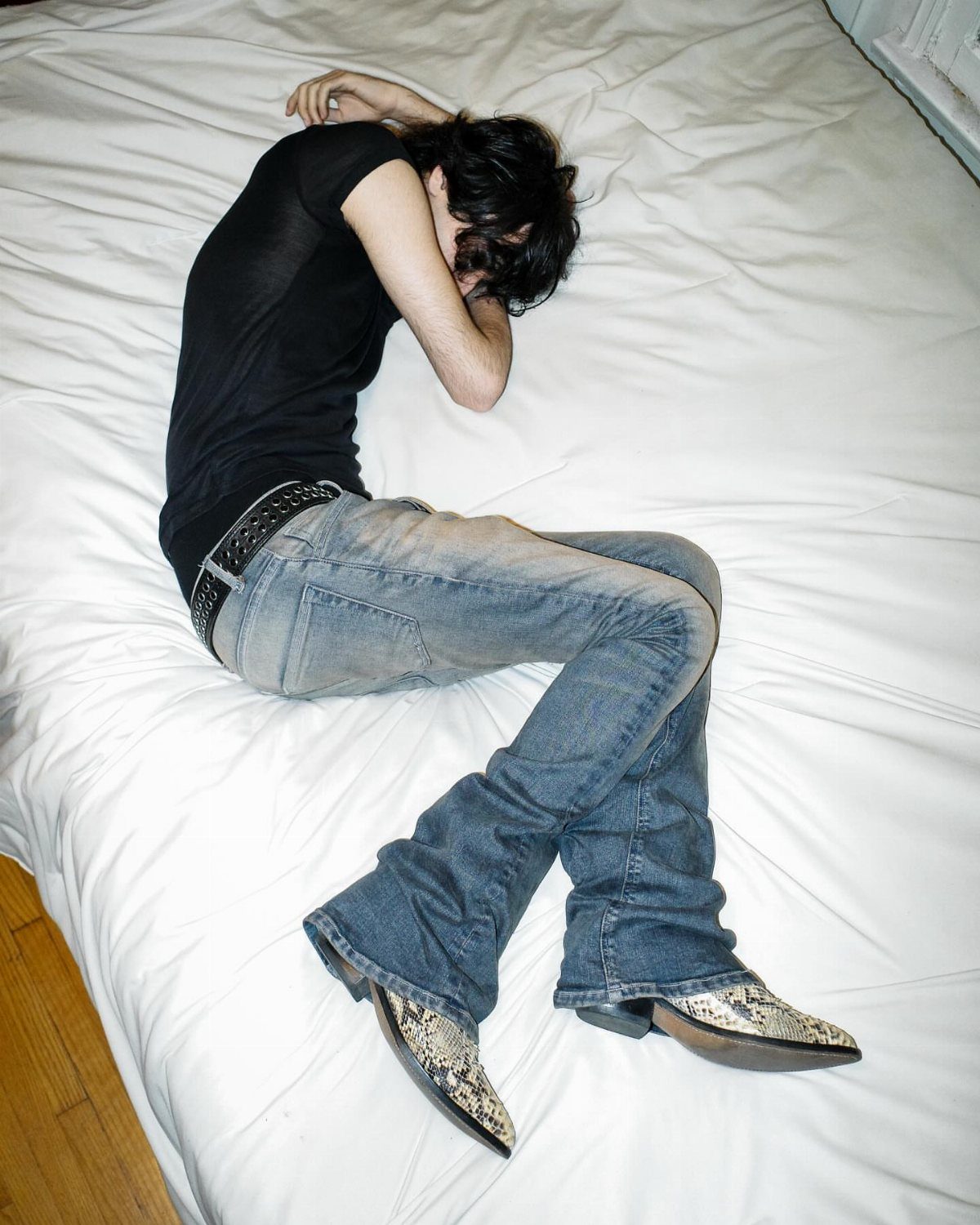 A person in black shirt, flared jeans, and snakeskin boots lying on a white bed.
