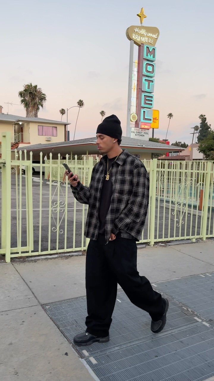 Man walking in baggy black pants, a plaid flannel shirt, and a black beanie outdoors.