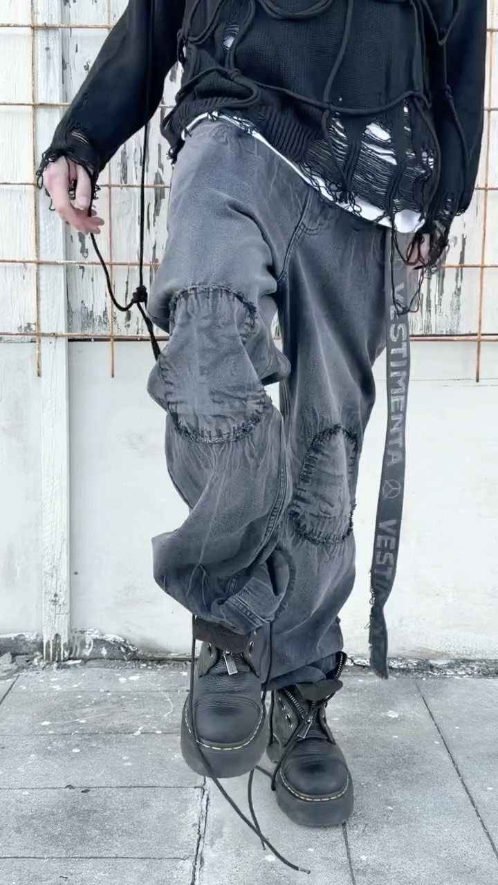 Person wearing oversized dark grey distressed denim trousers with large patches and black combat boots.