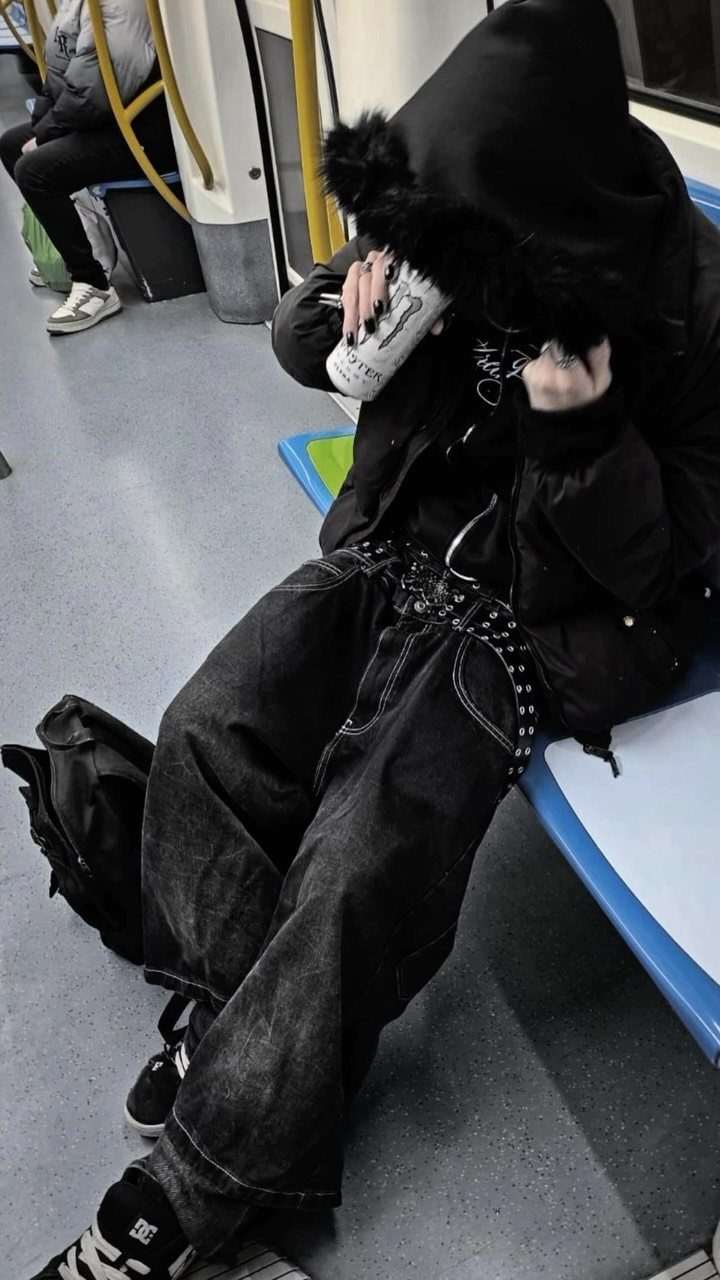 Person in black oversized hoodie and baggy studded denim pants sitting on a blue train seat.