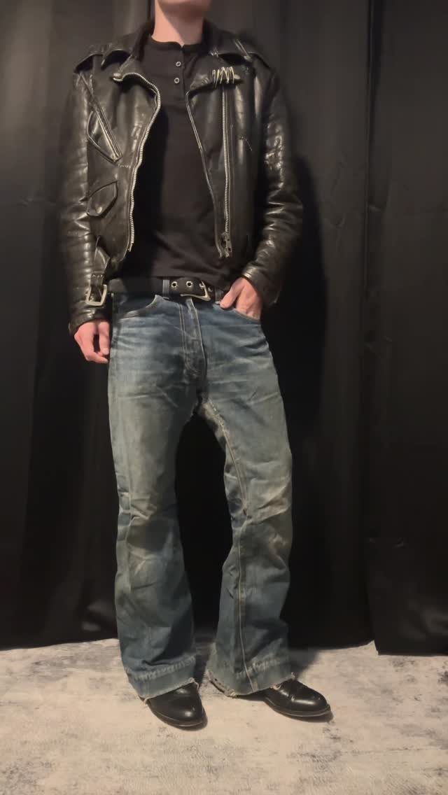 A person wearing a black leather jacket, black shirt, and faded blue jeans against a black background.