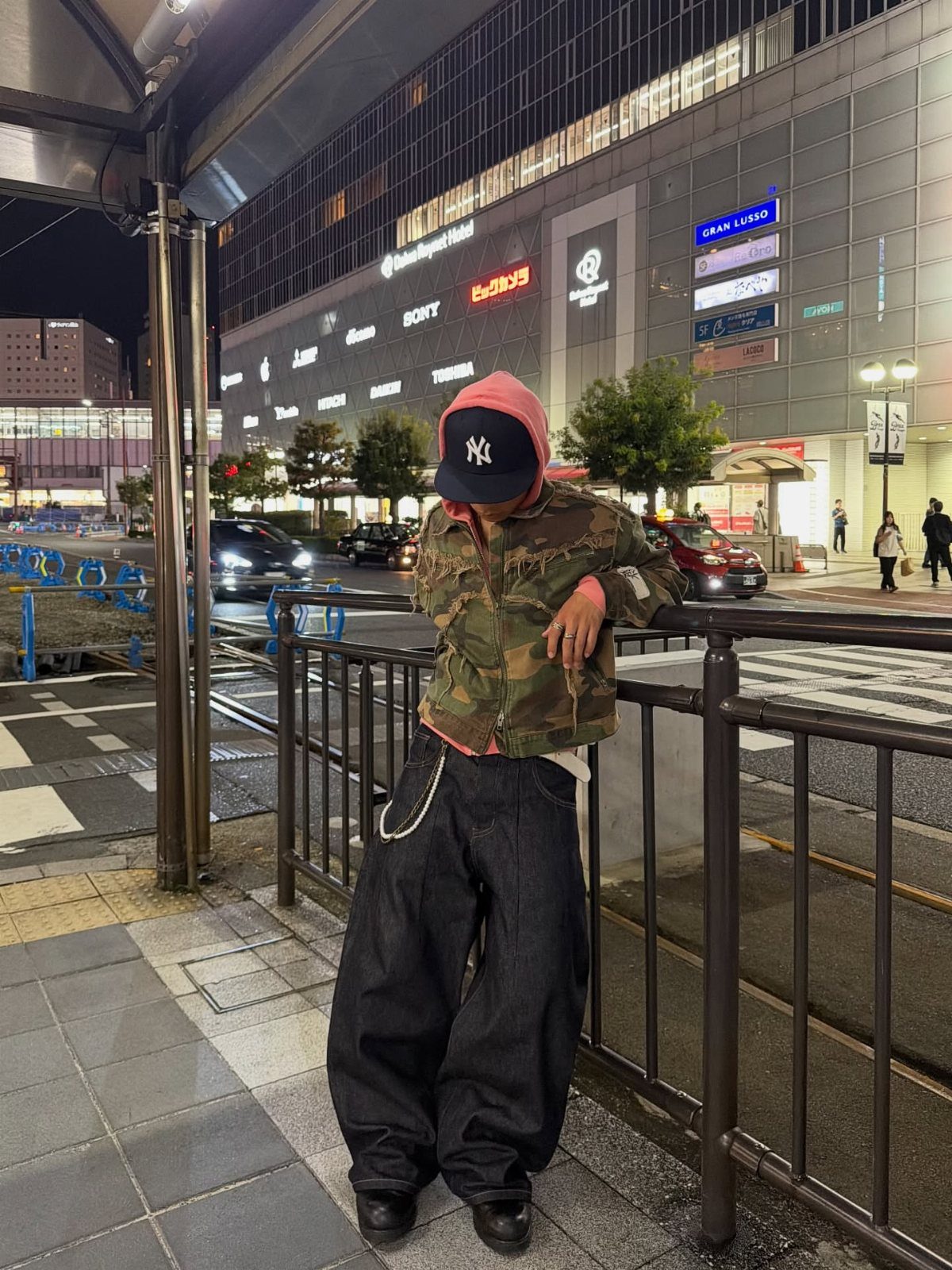 A person in camouflage and baggy black pants leans against a railing in a city at night.