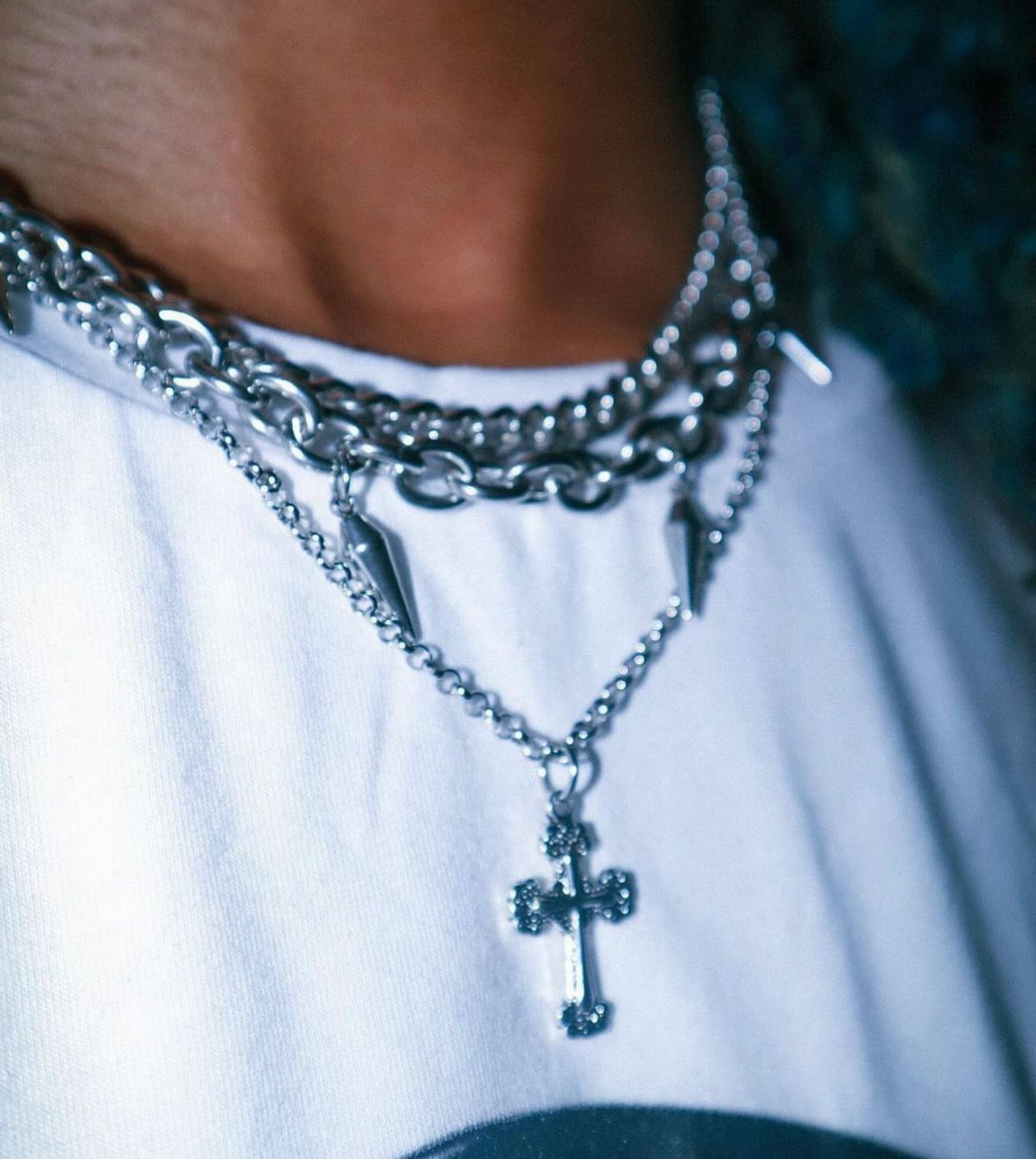 Silver chain necklaces with a cross pendant and spike charms worn over a white t-shirt.