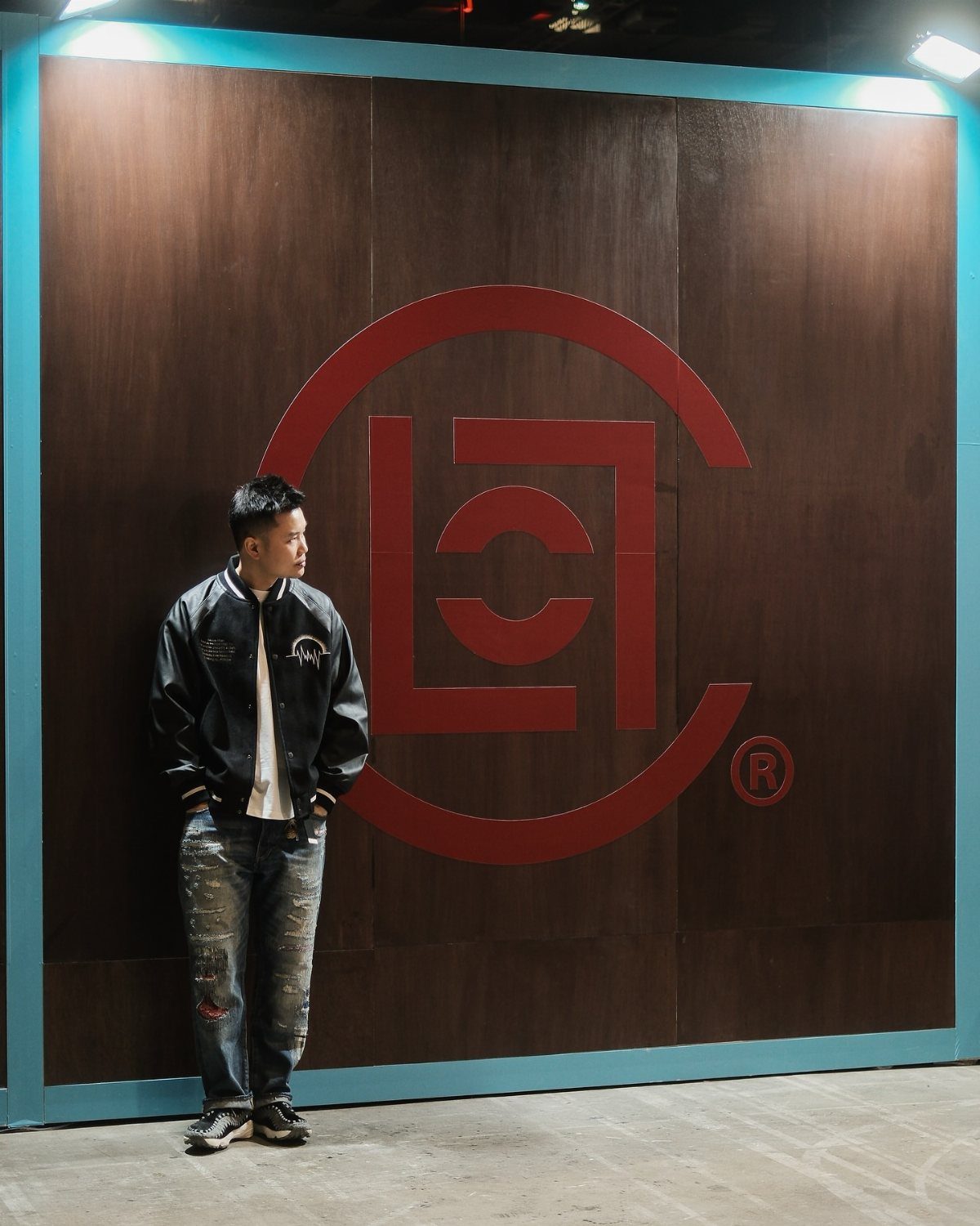 Man in streetwear standing before a large red circular logo on a dark wood wall.