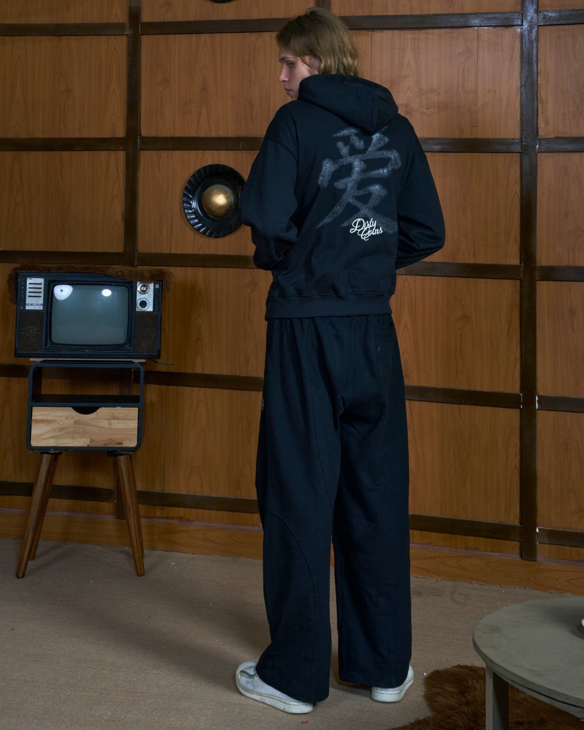 A person in black streetwear with kanji on the hoodie stands in a wood-paneled room.