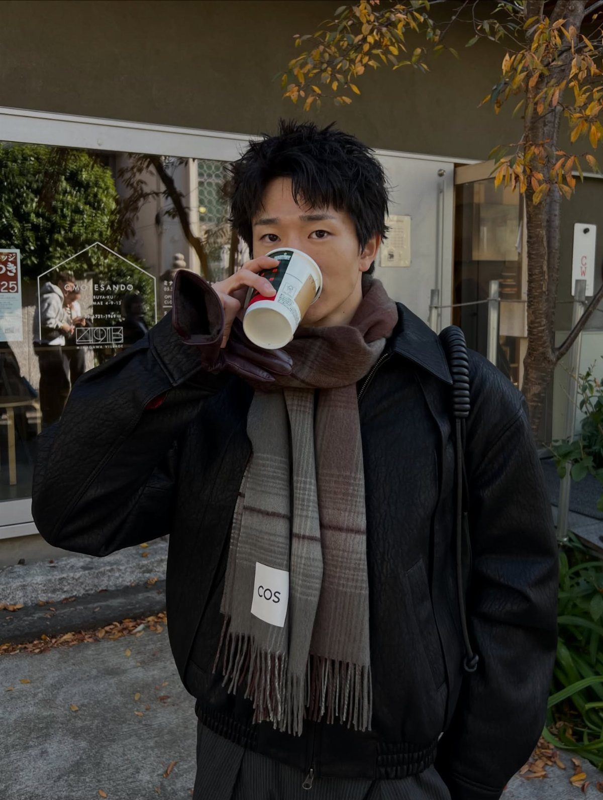 A young man in a black jacket and plaid scarf drinking from a coffee cup.
