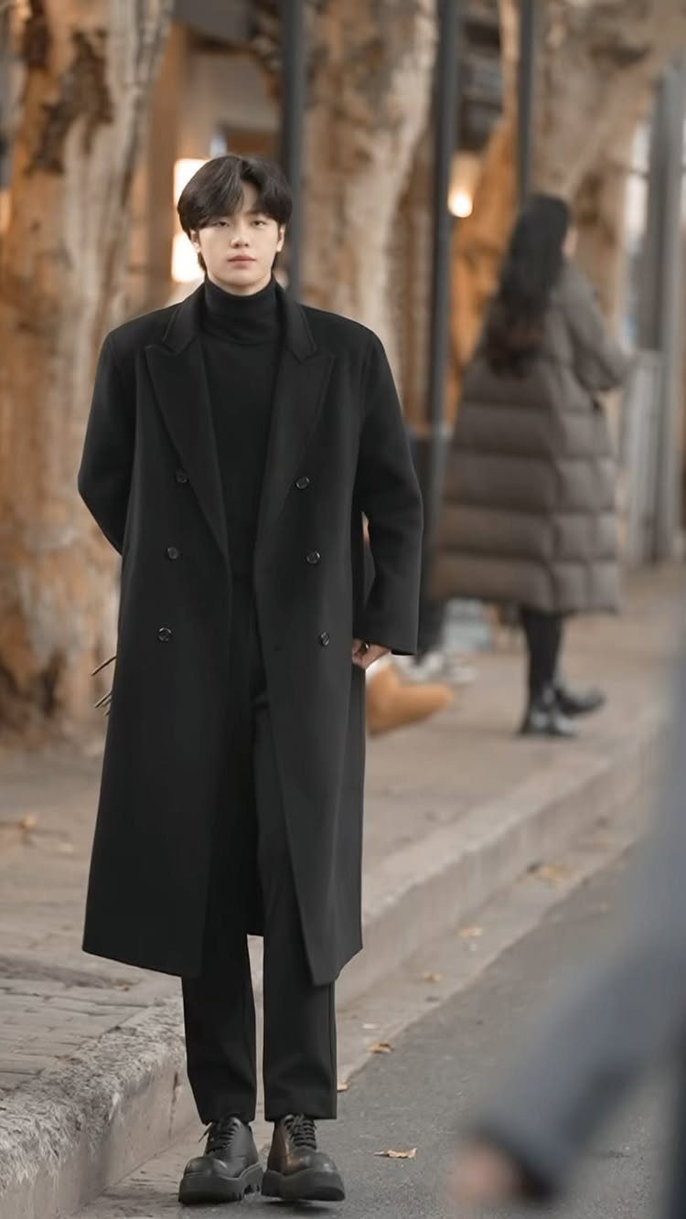 A man in a long black coat and turtleneck standing on a city sidewalk.