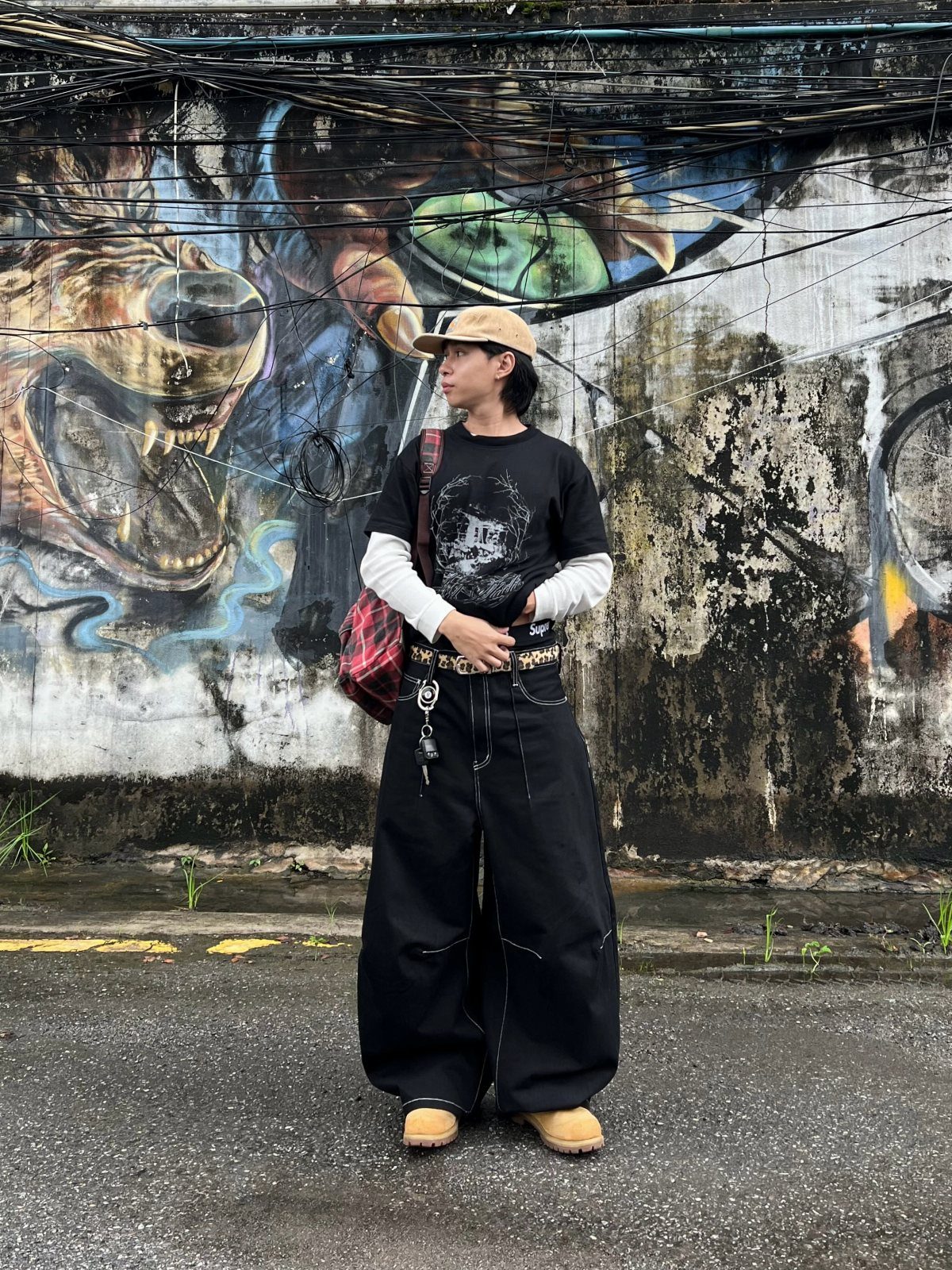 A person in graphic streetwear and wide-leg black trousers posing against a graffiti wall.