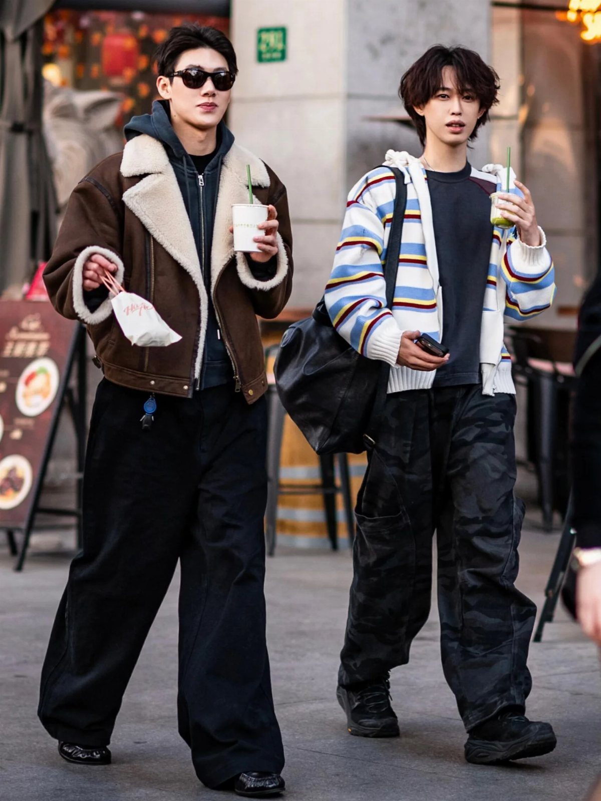 Two young men walking outdoors wearing oversized streetwear, including a shearling jacket and striped cardigan.