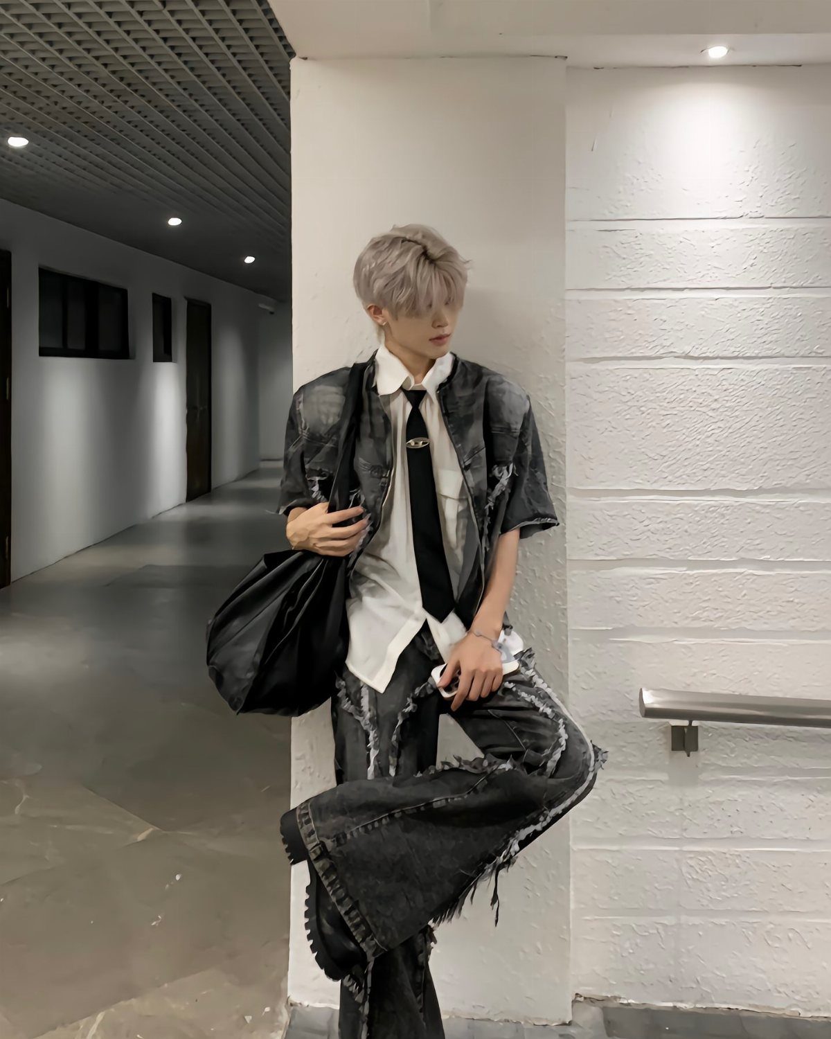 A person with silver hair wearing oversized black denim and a white shirt with a black tie.