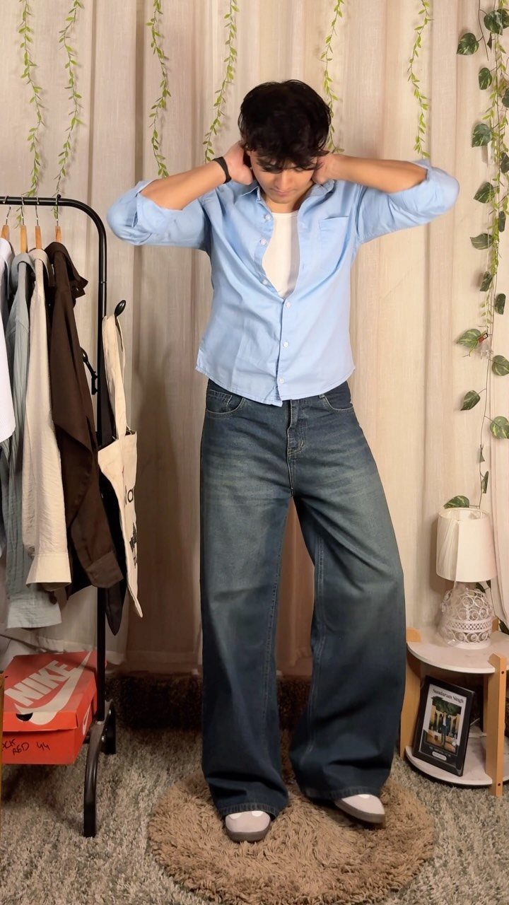 A man posing in a light blue button-down shirt and baggy blue denim jeans.
