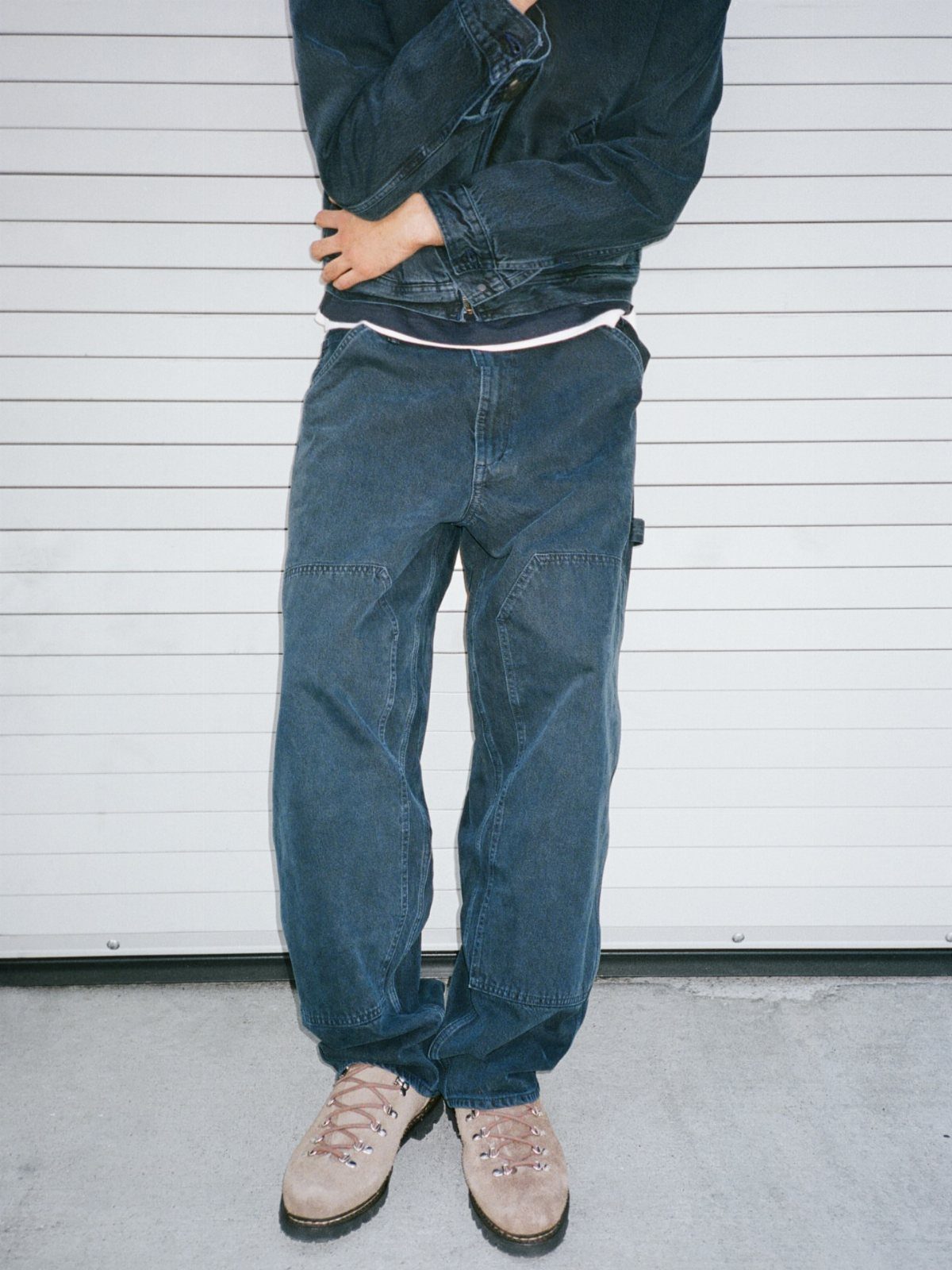 A person wearing baggy blue denim jeans, a dark denim jacket, and tan suede boots.