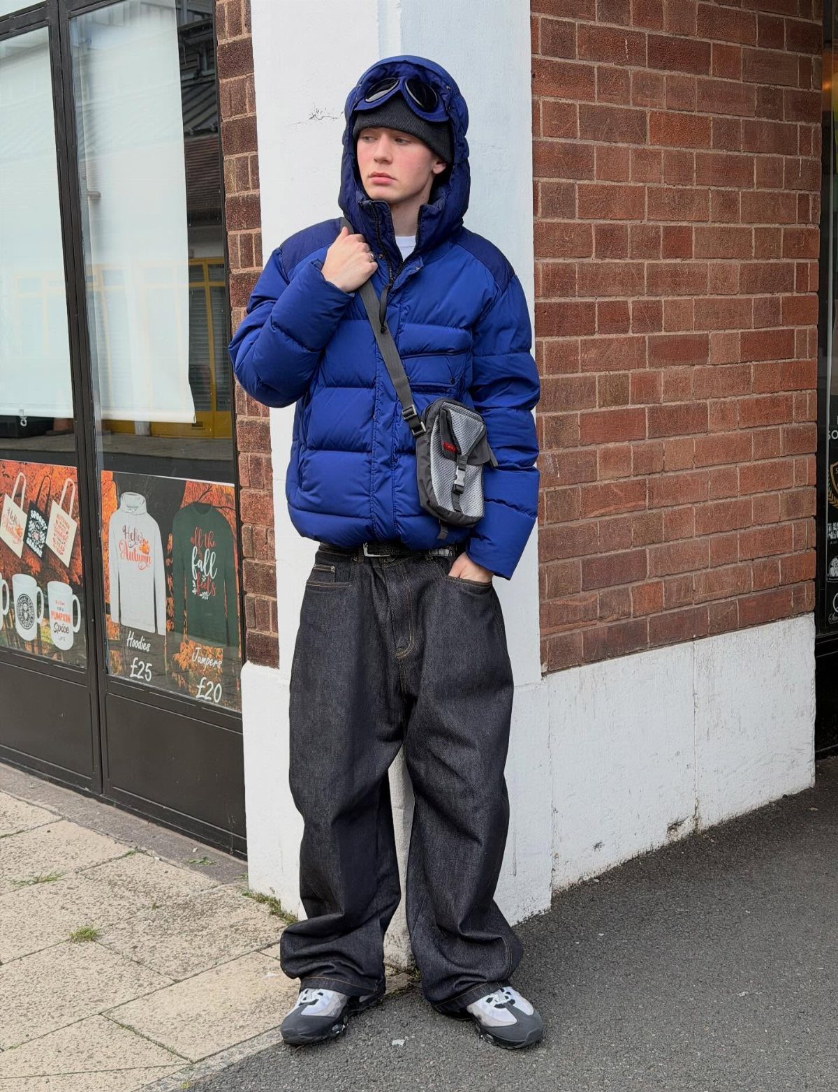 Man wearing a blue puffer jacket and baggy dark denim jeans standing by a brick wall.