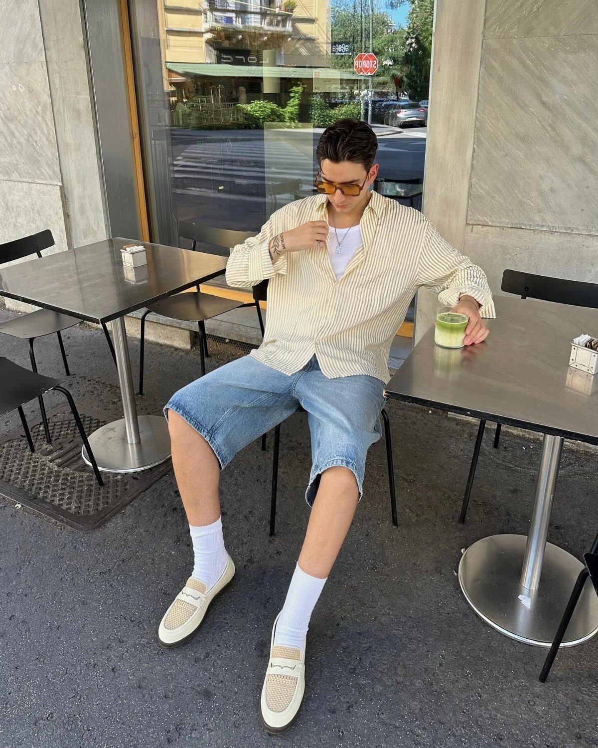 Man sitting at a cafe table wearing baggy denim shorts, a striped shirt, and loafers.