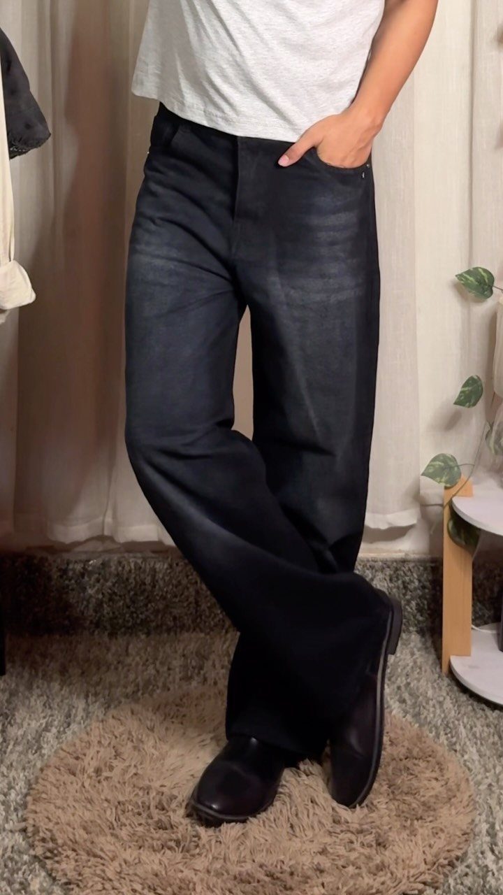 Person wearing dark baggy jeans, a white t-shirt, and black loafers standing on a rug.
