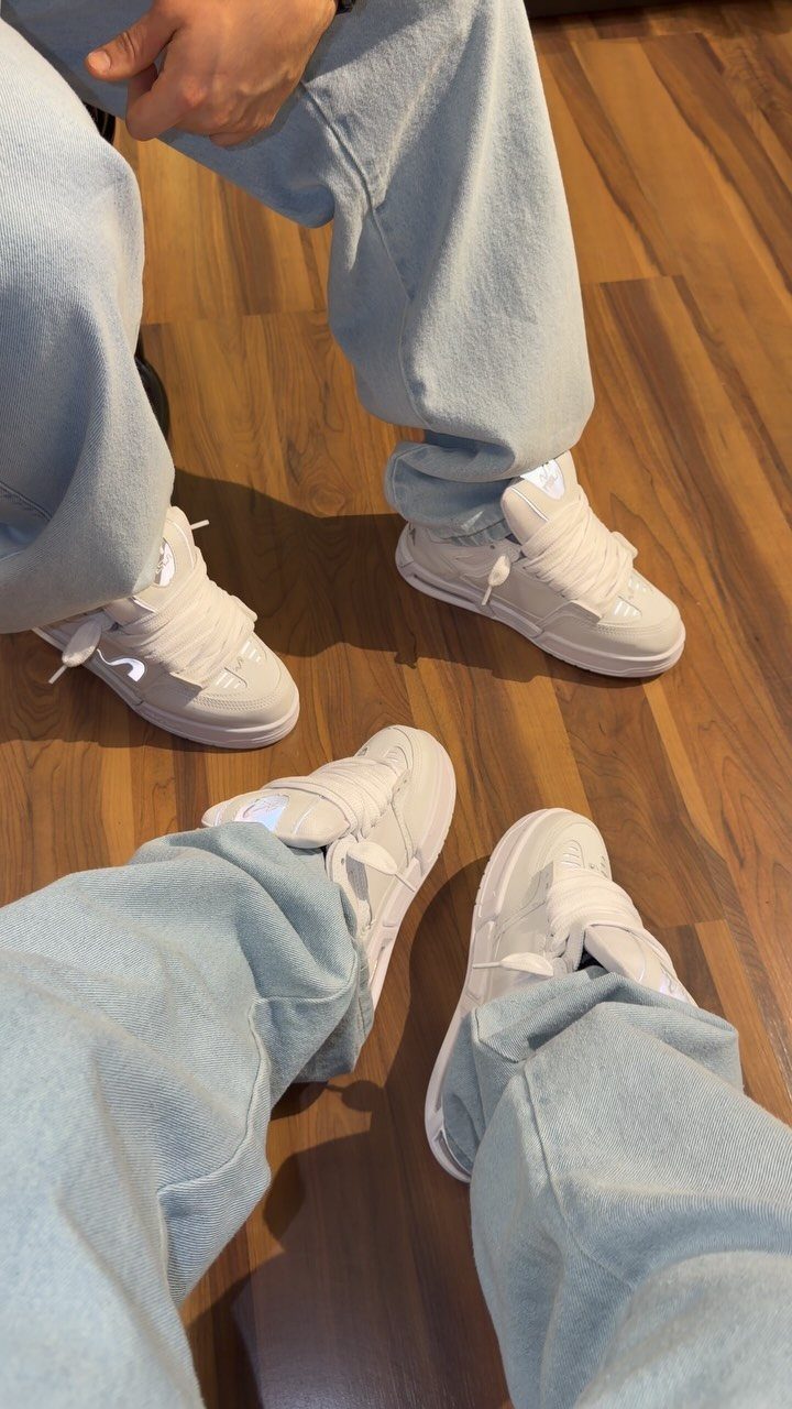 White chunky sneakers worn with light blue baggy jeans on a wooden floor.
